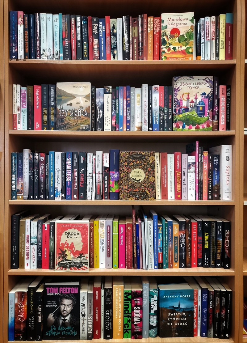 Drewniany regał z kilkoma półkami wypełnionymi książkami. Na półkach stoją różnorodne tytuły: powieści, reportaże, książki popularnonaukowe i dla dzieci, o kolorowych okładkach, fot. Biblioteka Miejska FB