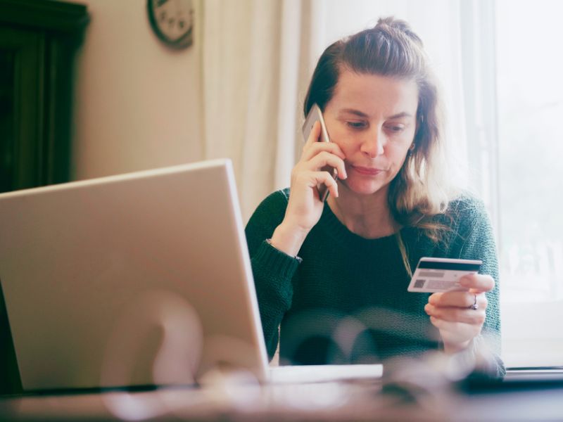 Kobieta w zielonym swetrze siedzi przy stoliku, rozmawiając przez telefon, trzymając w drugiej dłoni kartę bankomatową. Przed nią leży otwarty laptop. W tle widać okno z delikatnym światłem i zegar, tło jest rozmyte. fot. canva