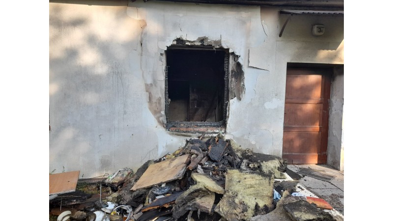 Zdjęcie okna lokalu, w którym wybuch pożarł, przedstawia białą ścianę z dziurą po oknie. Przed budynkiem znajdują się rzeczy ze spalonego mieszkania usunięte przez okno przez strażaków podczas akcji gaśniczej.