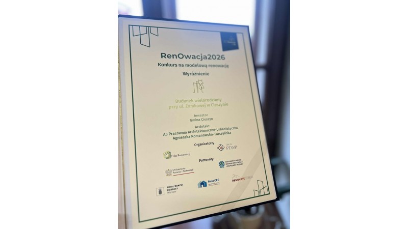 Zdjęcie przedstawia dyplom w jasnej ramce, ustawiony pionowo, na tle rozmytego wnętrza. Na dyplomie widoczny jest tytuł „RenOwacja2026” oraz informacja o konkursie na modelową renowację. Dokument przyznaje wyróżnienie dla budynku wielorodzinnego przy ul. 
