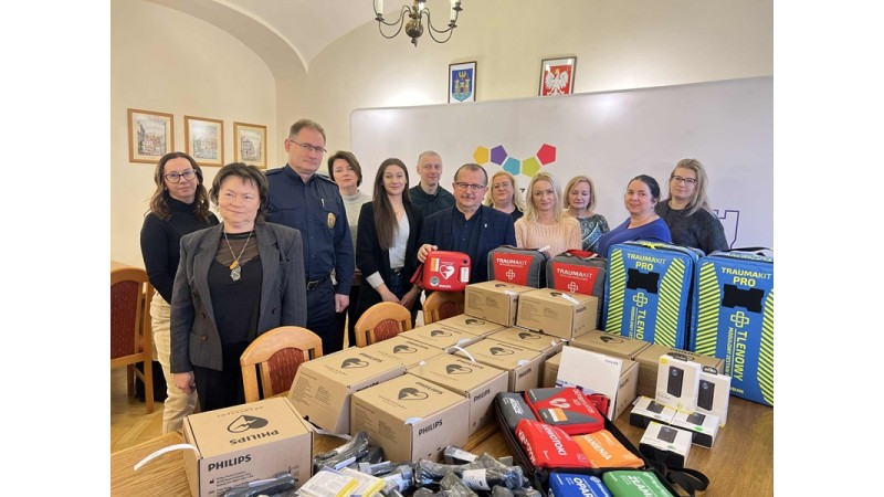 Grupa dorosłych osób stoi w sali urzędu za stołem z nowym sprzętem medycznym i ratowniczym, w tym defibrylatorami AED w kartonach oraz torbami i plecakami ratowniczymi. Zdjęcie przedstawia oficjalne przekazanie sprzętu na potrzeby ochrony ludności i dział