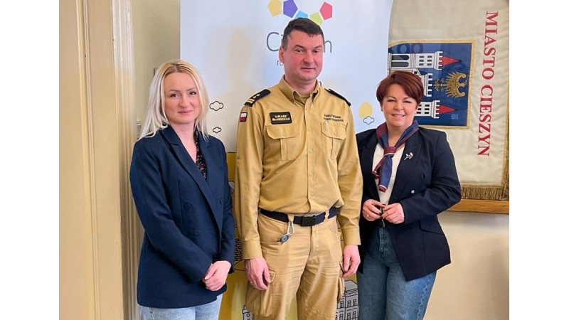 Zdjęcie przedstawia trzy osoby stojące obok siebie w przestrzeni urzędowej. W tle widoczny napis „Miasto Cieszyn”.